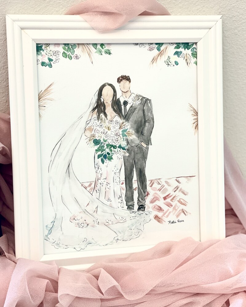 A framed watercolor portrait of a bride and groom, recreated from a photograph, resting on blush chiffon
