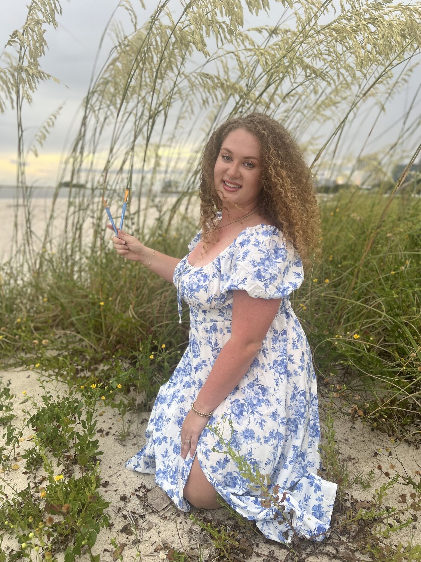 Katie on the beach in a blue and white dress, paintbrushes in hand