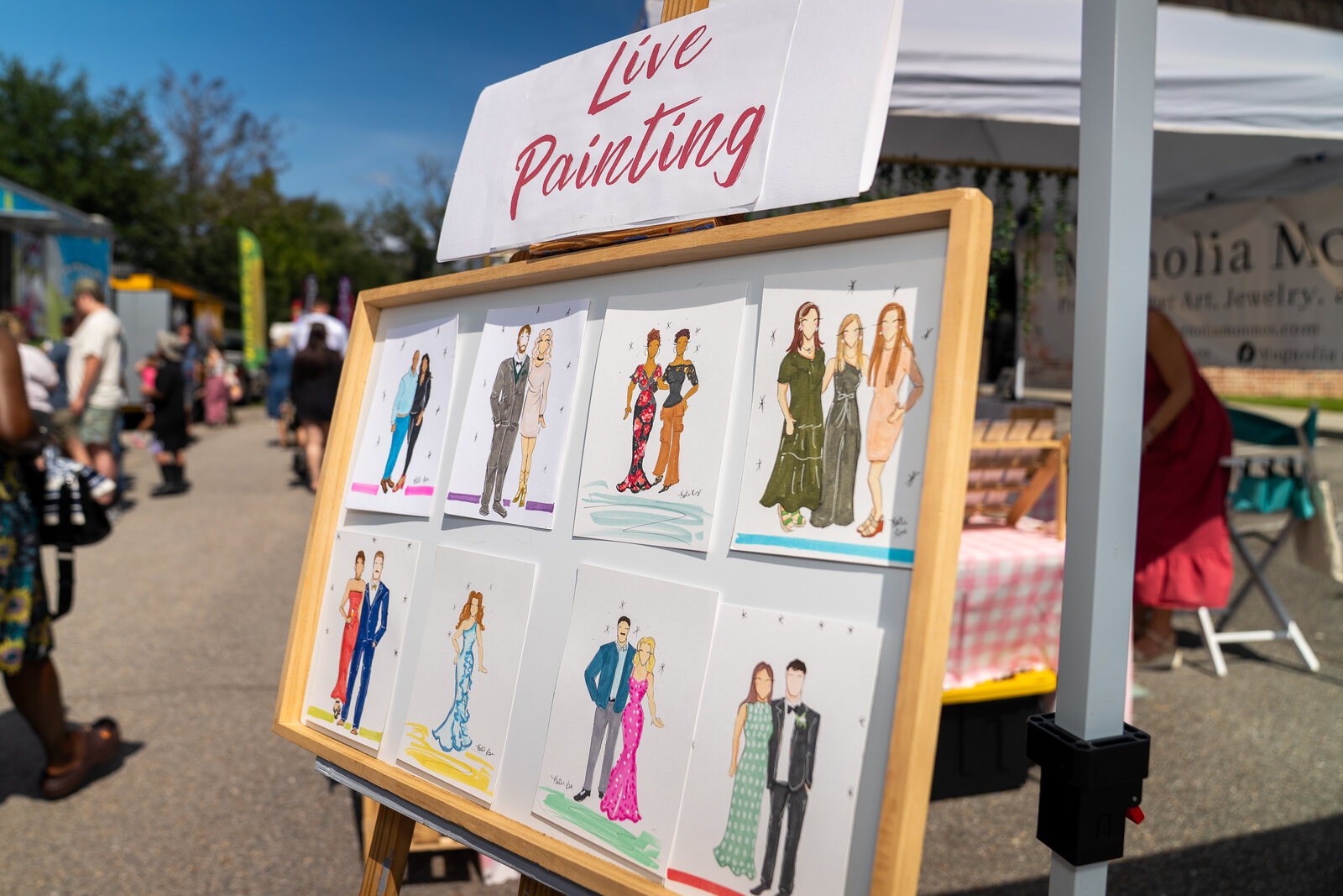 A board of Katie's fashion illustrations on an easel beneath a hand-lettered Live Painting sign at an outdoor event